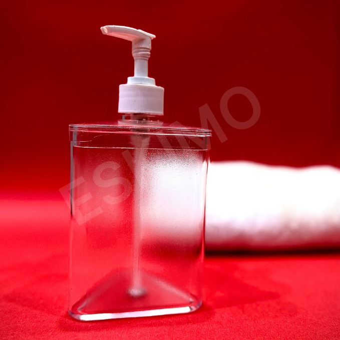 A transparent, triangular soap dispenser with a white pump head against a bright red background. A white towel is in the background.